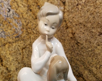 May include: A porcelain figurine of a young girl with a dog. The girl is wearing a white dress and has her finger to her lips. The dog is brown and white and is sitting next to the girl.