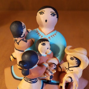 Puede incluir: Una figurita de cerámica pintada a mano de una madre y sus hijos. La madre lleva un vestido turquesa con un cinturón marrón y un collar. Tiene el pelo negro y los ojos marrones. Los niños llevan ropa marrón y amarilla. La figurita está hecha en un estilo tradicional.