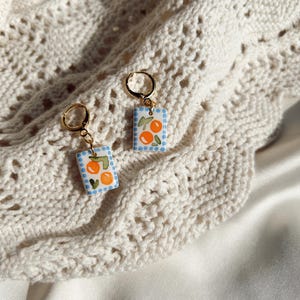 May include: A pair of gold hoop earrings with rectangular pendants. Each pendant features a hand-painted design of oranges and green leaves on a light blue background with white polka dots. The earrings are displayed on a cream-colored, open-knit fabric.