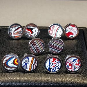 May include: A collection of round cufflinks with colorful, abstract designs. The cufflinks feature a variety of patterns in red, white, blue, and other colors, set in silver-toned metal. The cufflinks are displayed on a black, textured surface.