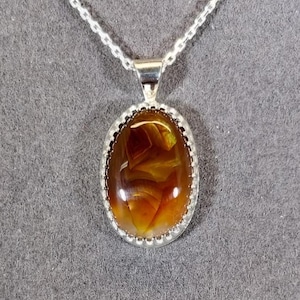 May include: A silver necklace featuring an oval pendant with a polished, amber-colored stone. The stone has swirling patterns of brown and yellow. The pendant is set in a silver bezel with a beaded edge, and hangs from a delicate silver chain.