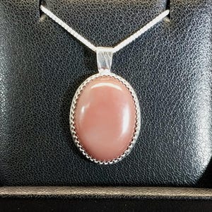 May include: A silver necklace featuring an oval-shaped, smooth, dusty rose-colored stone pendant. The pendant is set in a silver frame with a beaded edge and hangs from a thin silver chain, centered on a black textured display stand.