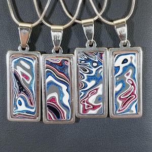 May include: Four silver pendant necklaces with rectangular, abstract-patterned pendants in shades of blue, white, gray, and red. The pendants are displayed on a black surface, with a wooden display stand in the background.