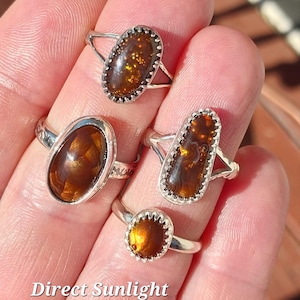 May include: Four silver rings with oval-shaped agate stones. The stones are a mix of brown, orange, and gold colors. The rings are displayed on a hand in direct sunlight outdoors. "Direct Sunlight Outdoors" is written below the rings.