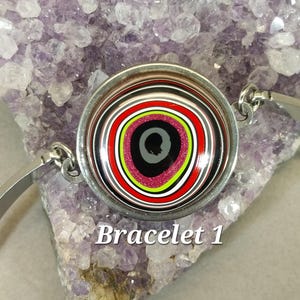 May include: A silver bracelet with a round, colorful, multi-layered, concentric circle design. The center of the circle is black with a pink and green ring around it. The bracelet is labeled "Bracelet 1".
