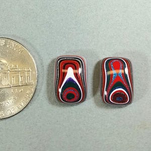 May include: Two rectangular, colorful cabochons with a swirled pattern of red, white, blue, and black. The cabochons are next to a U.S. nickel coin for size comparison.
