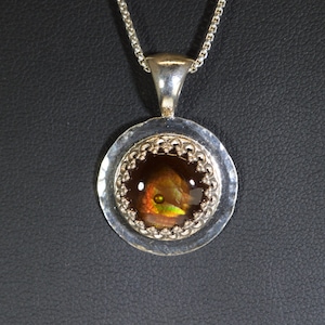 May include: A silver pendant necklace with a round, iridescent gemstone set in a silver bezel. The bezel has a decorative, scalloped edge.