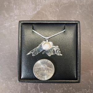 May include: A silver-toned pendant shaped like the outline of Lake Superior, featuring a small circular white stone set in the center. The pendant hangs from a thin silver chain and is displayed next to a United States five-cent nickel coin.