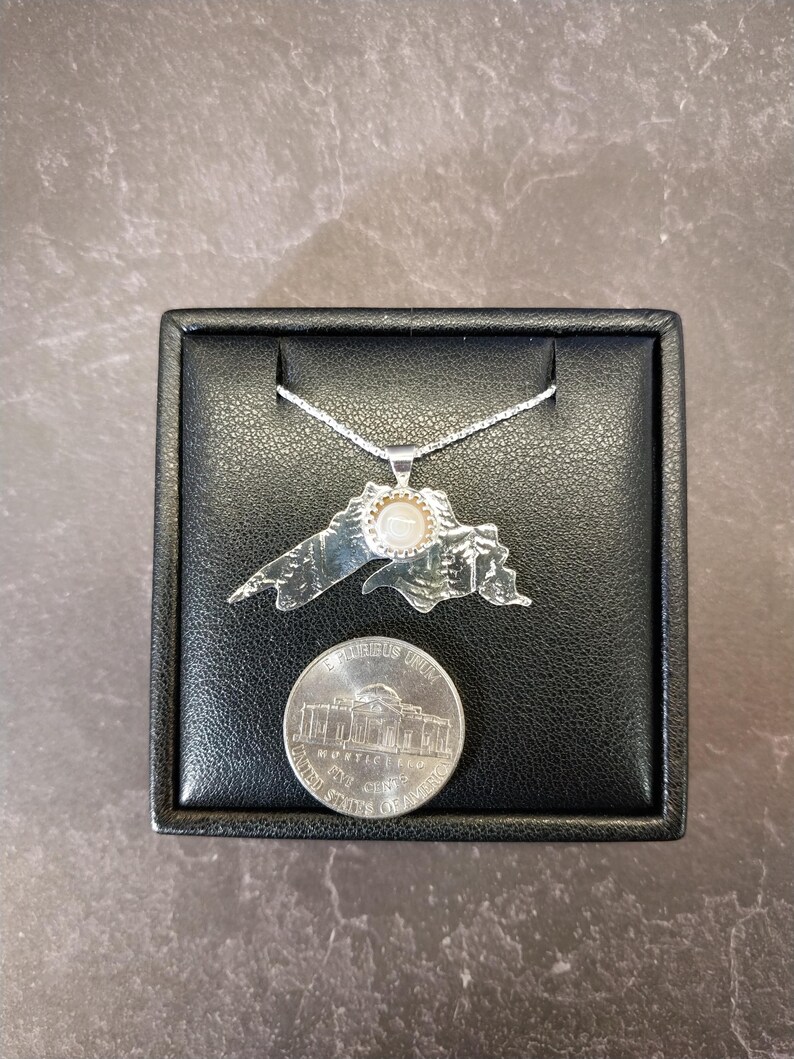 May include: A silver-toned pendant shaped like the outline of Lake Superior, featuring a small circular white stone set in the center. The pendant hangs from a thin silver chain and is displayed next to a United States five-cent nickel coin.