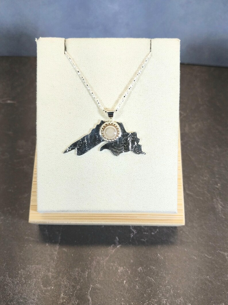 May include: A silver-toned pendant necklace featuring a textured, map-shaped charm with a jagged outline. A small, circular, light-colored stone is set in a serrated bezel at the top center of the pendant, which hangs from a delicate silver chain.