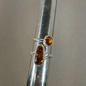 May include: Two silver rings with amber stones. The ring on the bottom has a rectangular stone, while the ring on top has a round stone.
