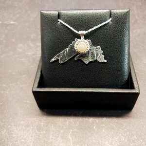 May include: A silver pendant shaped like the outline of Lake Superior, featuring a textured metallic surface and a small, round, light-colored stone set in the center. The pendant hangs from a thin silver chain.