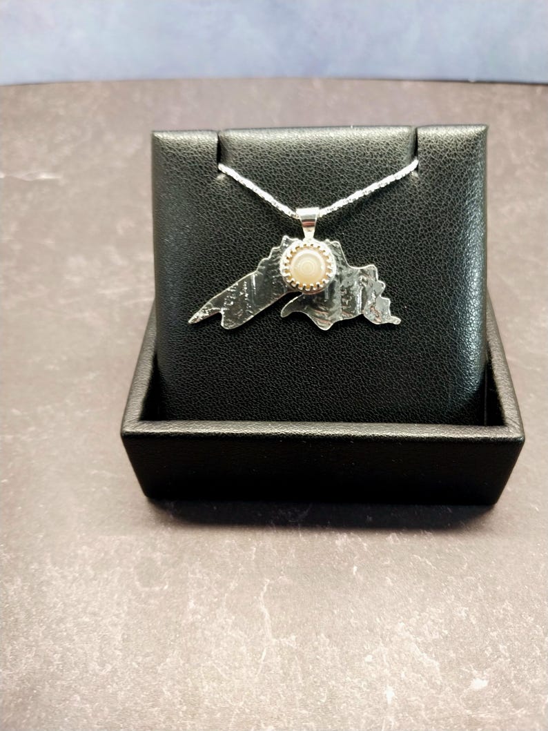 May include: A silver pendant shaped like the outline of Lake Superior, featuring a textured metallic surface and a small, round, light-colored stone set in the center. The pendant hangs from a thin silver chain.