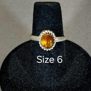 May include: A silver ring with a round, orange gemstone set in a beaded bezel. The ring is on a black ring display stand. Size 6.