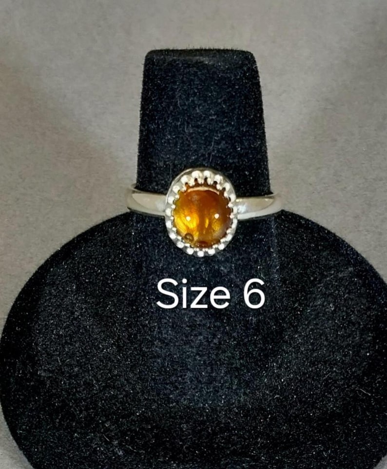 May include: A silver ring with a round, orange gemstone set in a beaded bezel. The ring is on a black ring display stand. Size 6.