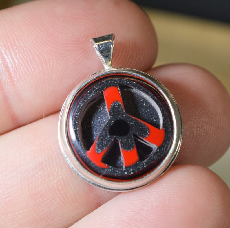 May include: A silver pendant with a red and black peace sign inside a glass dome.