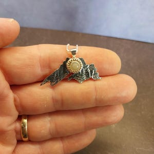 May include: A silver pendant shaped like the outline of Lake Superior with a textured, oxidized finish. A small, round, light-colored stone is bezel-set in gold at the center of the pendant, which hangs from a delicate silver chain.