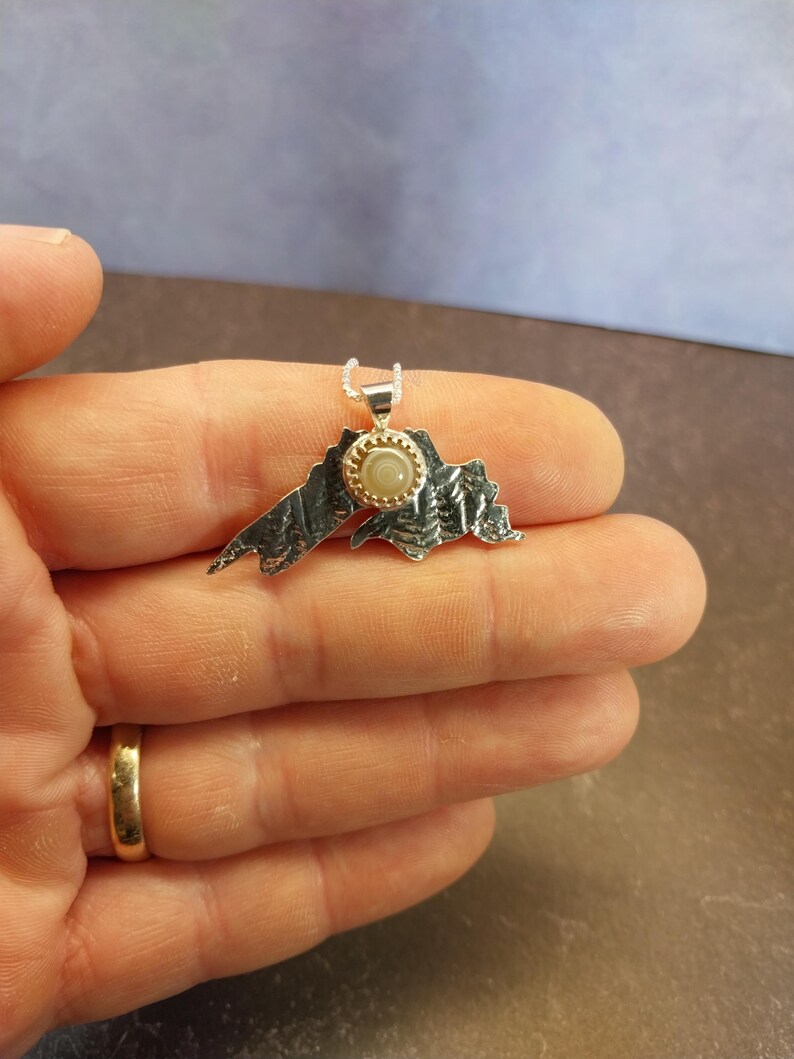 May include: A silver pendant shaped like the outline of Lake Superior with a textured, oxidized finish. A small, round, light-colored stone is bezel-set in gold at the center of the pendant, which hangs from a delicate silver chain.