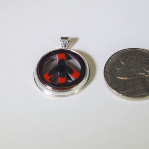 May include: A silver pendant with a peace sign design. The peace sign is black and red with a silver border. The pendant is about the size of a US quarter.