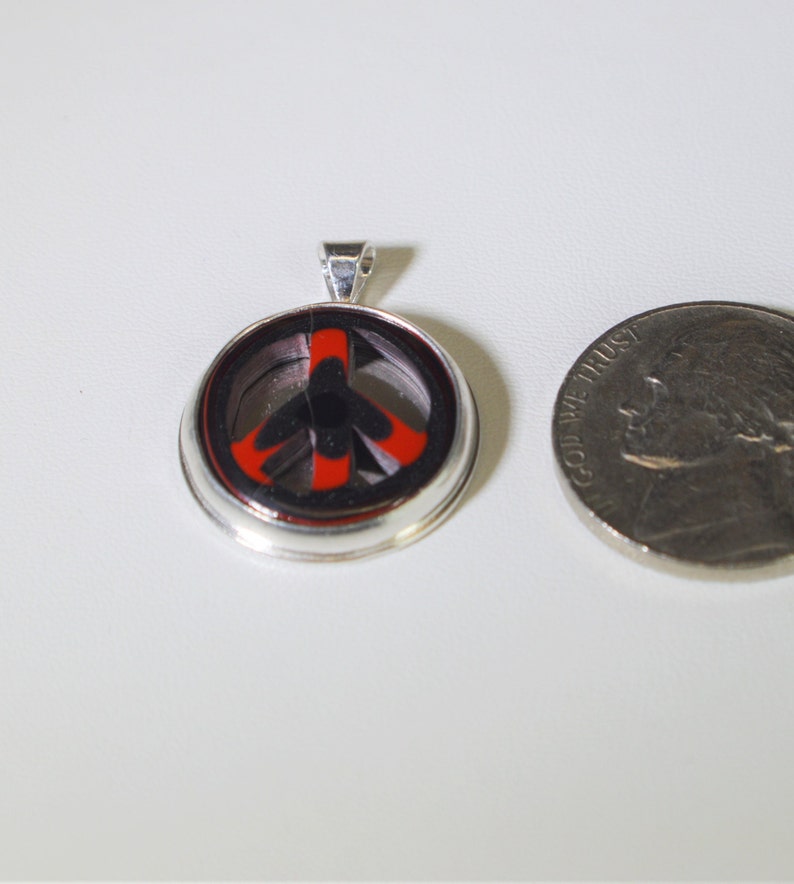 May include: A silver pendant with a peace sign design. The peace sign is black and red with a silver border. The pendant is about the size of a US quarter.