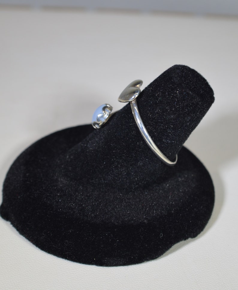 May include: A silver ring with a heart-shaped design and a blue stone. The ring is displayed on a black velvet ring holder.