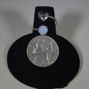 May include: Three silver rings on a black display stand. The top ring has a heart-shaped stone. The middle ring has a round, light blue stone. The bottom ring is a plain band.