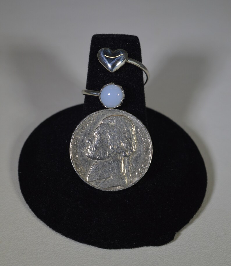 May include: Three silver rings on a black display stand. The top ring has a heart-shaped stone. The middle ring has a round, light blue stone. The bottom ring is a plain band.