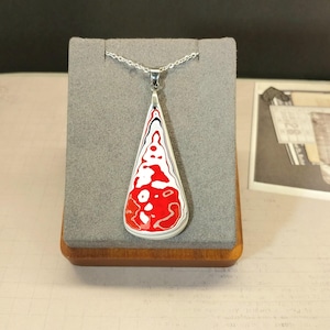 May include: A silver pendant necklace with a teardrop-shaped pendant. The pendant features a red, white, and black abstract design. The necklace is displayed on a gray stand with a wooden base.