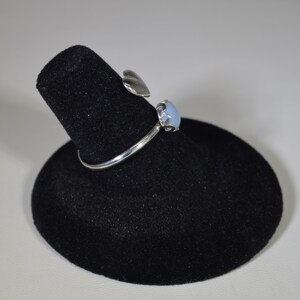 May include: A silver ring featuring a light blue gemstone and a heart-shaped detail. The ring is presented on a black velvet ring stand.
