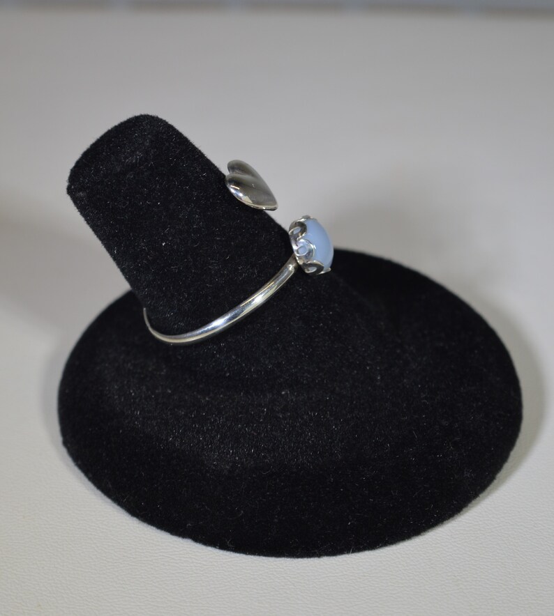 May include: A silver ring featuring a light blue gemstone and a heart-shaped detail. The ring is presented on a black velvet ring stand.
