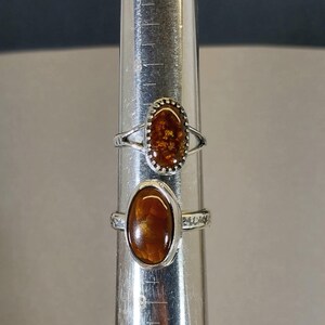 May include: Two silver rings with oval-shaped brown and orange gemstone settings. The rings are on a silver measuring stick.