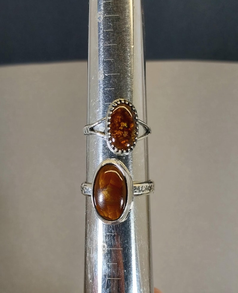 May include: Two silver rings with oval-shaped brown and orange gemstone settings. The rings are on a silver measuring stick.