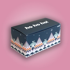 Puede incluir: Una caja de regalo azul y blanca con un patrón de montaña festivo y el texto "ho ho ho!" en la parte superior. La caja está hecha de cartón y tiene un logotipo YAWN blanco en el lateral.