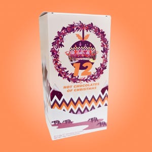 May include: A white box with a Christmas theme, featuring a purple wreath and a decorative ornament with the number 12. The text "HOT CHOCOLATES OF CHRISTMAS" is displayed below the ornament. The box has a zig-zag pattern in orange and purple.