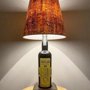 May include: A table lamp made from a Colonel E.H. Taylor whiskey bottle. The lamp features a brown lampshade with a textured pattern and a square base. The bottle's label is visible, with text in yellow and black.
