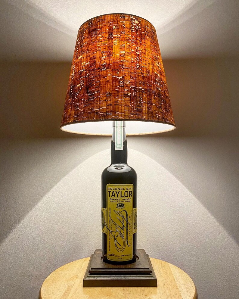 May include: A table lamp made from a Colonel E.H. Taylor whiskey bottle. The lamp features a brown lampshade with a textured pattern and a square base. The bottle's label is visible, with text in yellow and black.
