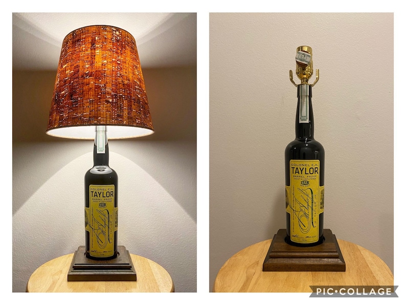 May include: A unique table lamp crafted from a Colonel E.H. Taylor whiskey bottle. The lamp features a dark brown lampshade with a textured pattern and a wooden base. The bottle has a yellow label with black text.