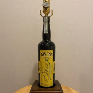 May include: A lamp made from a Colonel E.H. Taylor bourbon whiskey bottle. The black bottle has a gold label with text and is mounted on a wooden base. The lamp has a gold-colored socket and a small piece of paper attached to it.
