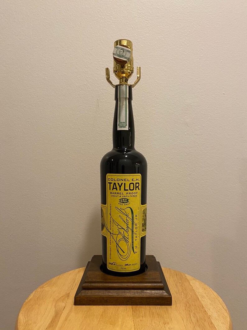 May include: A lamp made from a Colonel E.H. Taylor bourbon whiskey bottle. The black bottle has a gold label with text and is mounted on a wooden base. The lamp has a gold-colored socket and a small piece of paper attached to it.