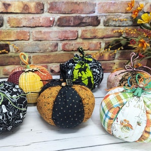 May include: Five handmade fabric pumpkins in various patterns and colors. The pumpkins are decorated with black and white ghost patterns, plaid, and a green and black pattern. The pumpkins are sitting on a white wooden surface.