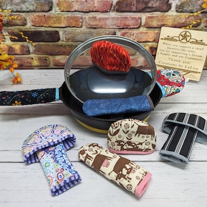 Puede incluir: Un conjunto de cinco agarraderas de olla coloridas, que incluyen una agarradera de olla a rayas blancas y negras, una agarradera de olla floral, una agarradera de olla con temática de vaca, una agarradera de olla con diseño de hongo y una agarradera de olla con diseño de calavera y flor. Las agarraderas de olla se muestran sobre una superficie de madera blanca con una agarradera de olla con estampado blanco y negro sobre una sartén negra.