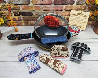 May include: A black skillet with a glass lid and several fabric handle covers in various patterns including floral, cow print, skull, and striped designs. A small card with the text "When you support a small business like mine, you are supporting a Dream, a Family, and our Community. Thank you! Creations by LaLita" is visible.
