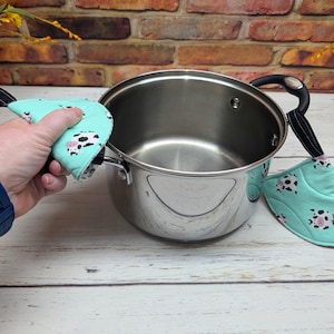 May include: A stainless steel cooking pot with two black handles, accompanied by two light mint green fabric handle covers featuring a repeating pattern of black and white cows with pink snouts, each with a black fabric hanging loop attached.