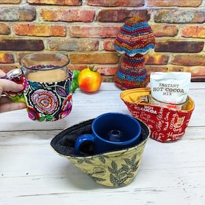 May include: Three fabric mug cosies in different patterns: one black with colourful floral prints, one red with white text reading Instant Hot Cocoa Mix, and one beige with black leaf motifs. Each cosy holds a mug, with one containing hot cocoa mix.