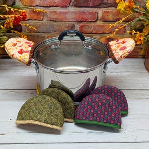 May include: A stainless steel pot with a glass lid and two matching pot holders. The pot holders have a mushroom design. Several pot handle covers in green and burgundy floral patterns are in front of the pot.