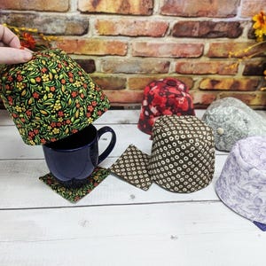 May include: A dark blue mug with a matching coaster is paired with several fabric cup cozies. The cozies feature various patterns, including floral, geometric, and abstract designs. The cozies are in shades of green, red, brown, and purple.