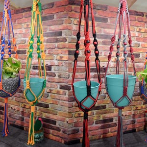 May include: Five macrame plant hangers in various colour combinations including blue, orange, green, yellow, black, red, and grey. Each hanger features knotted cord patterns, wooden beads, and a tassel at the bottom, holding either a solid or speckled pot.