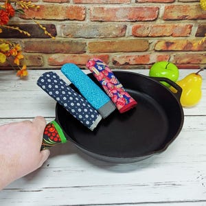 May include: A black cast iron skillet with three colorful handle covers. The handle covers feature patterns of white dots on navy, blue geometric shapes, and red strawberries. The skillet is on a white wooden surface.