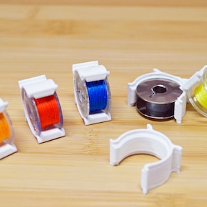 May include: Five white plastic bobbins for thread spools. Four bobbins are filled with thread in orange, blue, yellow, and a lighter orange. One bobbin is empty.
