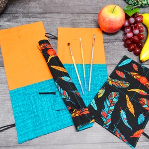 May include: Two fabric pencil cases with a black background and colourful feather pattern. The cases are rolled up and have a button closure. One case is open and has four paintbrushes inside. The cases are on a wooden surface with a banana, grapes, and an apple.